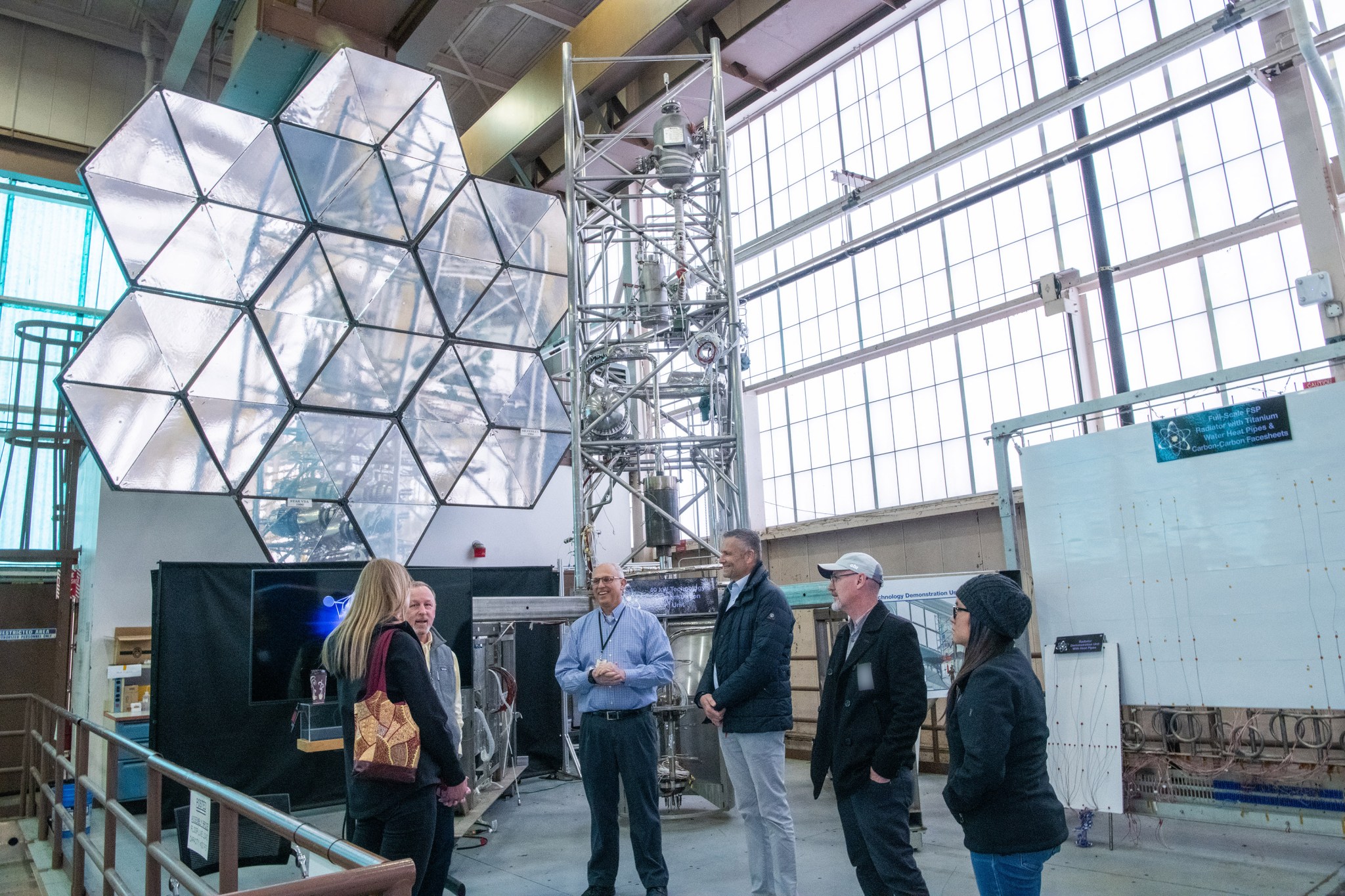 Group of six people stand in semicircle talking to one another. Scaffolding and large space mirror in background.