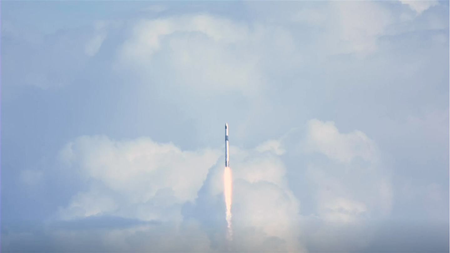 Most of the image is filled with a light blue sky and large, white and gray billowing clouds. Small, in the middle of the image, a white rocket launches, with a plume of fire and smoke below it.