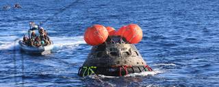 NASA’s Orion spacecraft for the Artemis I mission in the water with four orange round flotation devices on top of it. A boat is in the background.