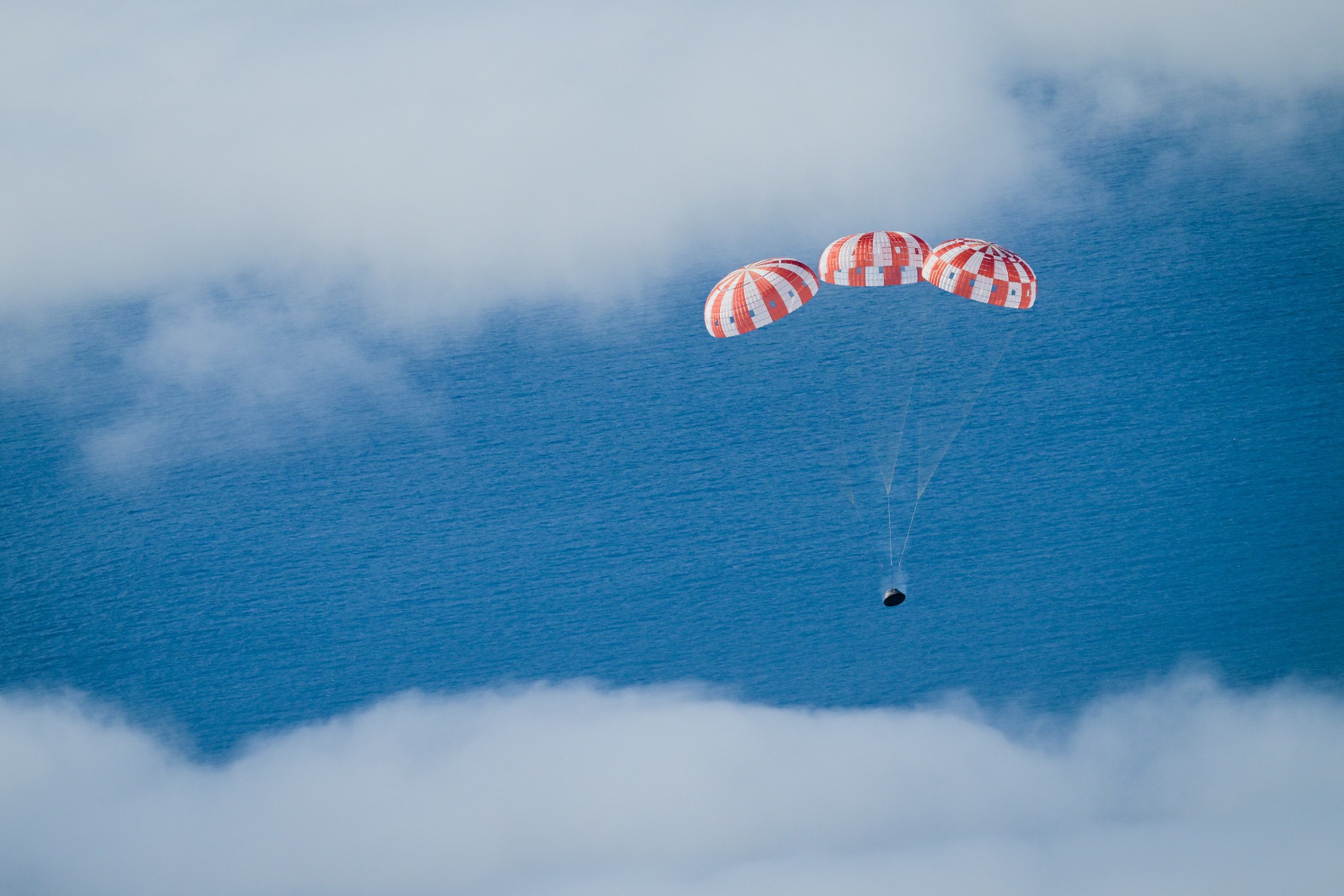 The Orion crew capsule with its three red and white parachutes is seen during the Artemis I mission descending through the clouds to the ocean surface, visible below.