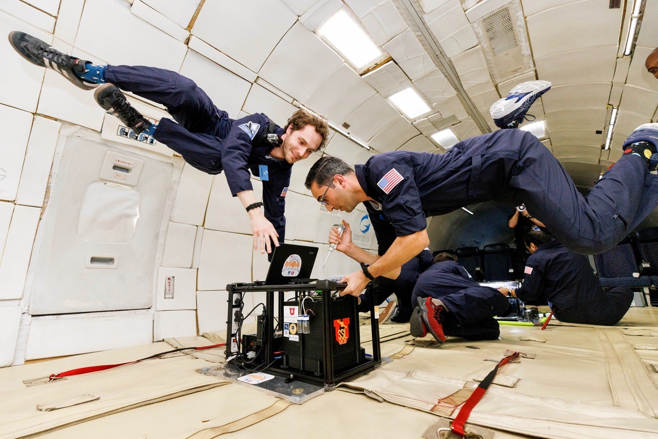 Inside the cabin of an airplane, two researchers in flight suits float in zero gravity. In front of them is a square black technology payload about the size of a microwave.