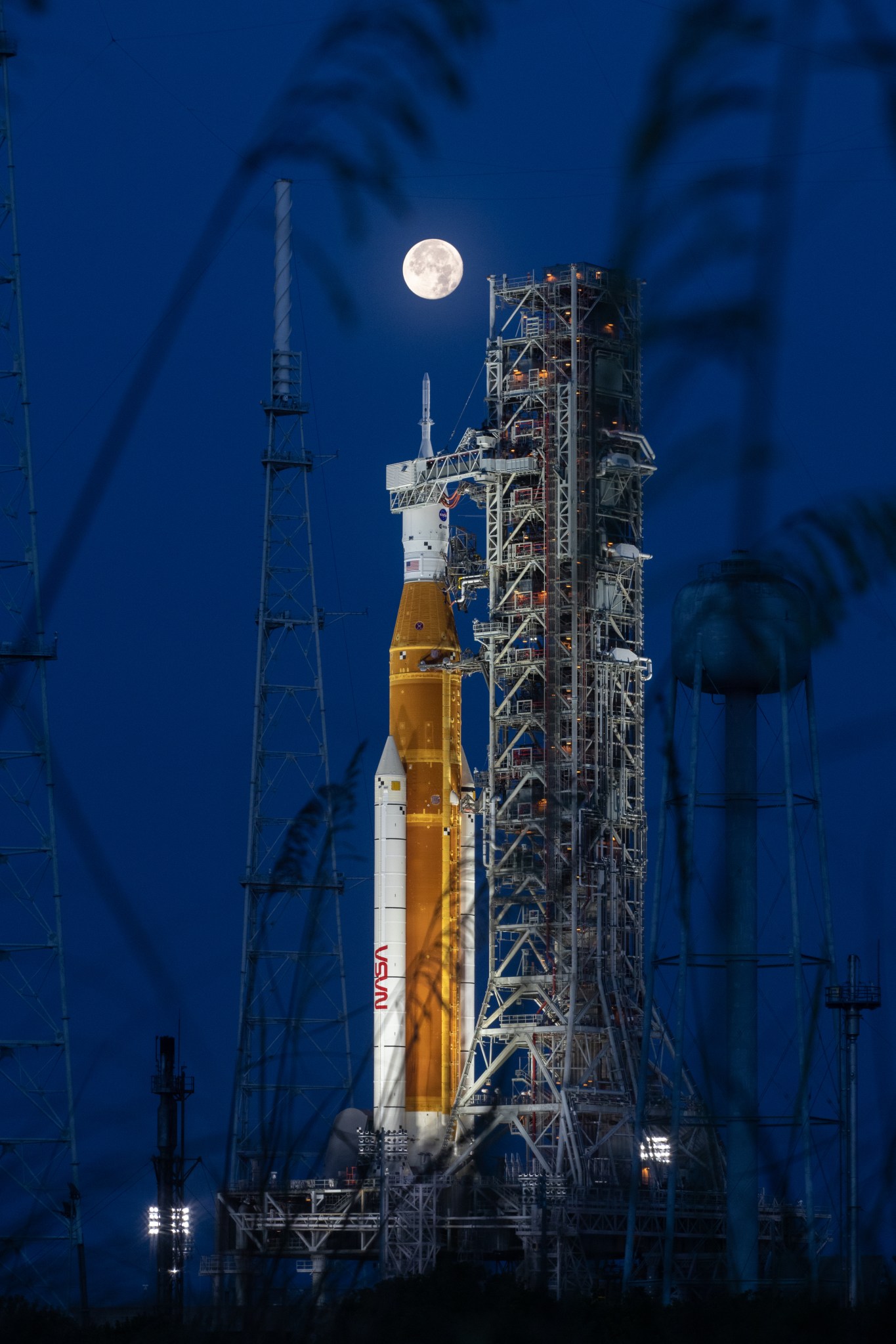 A full Moon can be seen above the Space Launch System rocket, poised at the launch pad at night. 