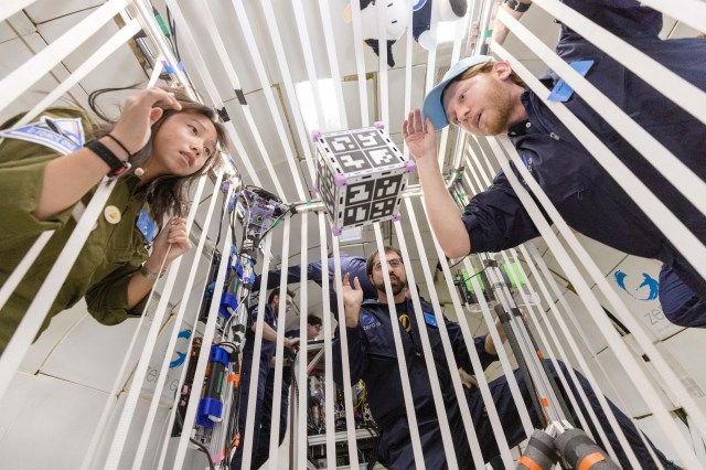 Three researchers peer through the white bands that create a cell-like space containing a floating cube with QR codes on each side. A camera mounted to the top corner of the cell records the experiment.