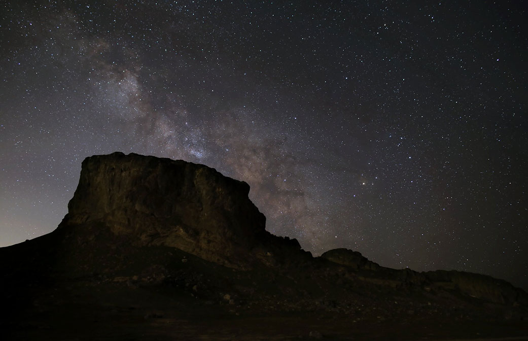 A photograph shows a desert butte in front of a night sky glittering with stars. The band of the Milky Way crosses the sky diagonally. It looks like a glowing cloud filled with dust and stars above the desert landscape.