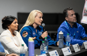 Three people sitting on a table with name tags on the table, while one of them talks in the microphone.