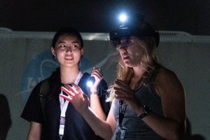 Two people in the dark. One person holding a flash light while the other person has their hands raised with AR goggles on.