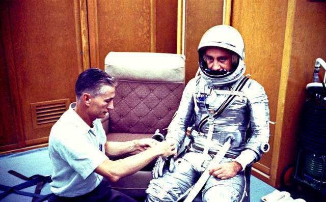 Gus Grissom waits in his space suit in the personal equipment room of Hangar S while spaceflight equipment specialist Joe W. Schmidt adjusts his glove.