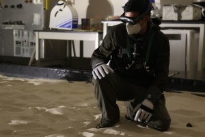 A person with goggles and mask is kneeling in a room with sand.
