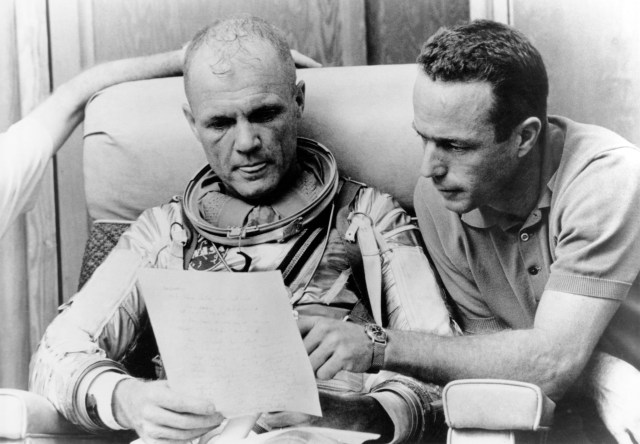 John Glenn, wearing his space suit, goes over notes on a piece of paper with backup pilot Scott Carpenter.