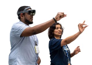 Two people standing and one arm raised with pinch sign. One person wears goggles.