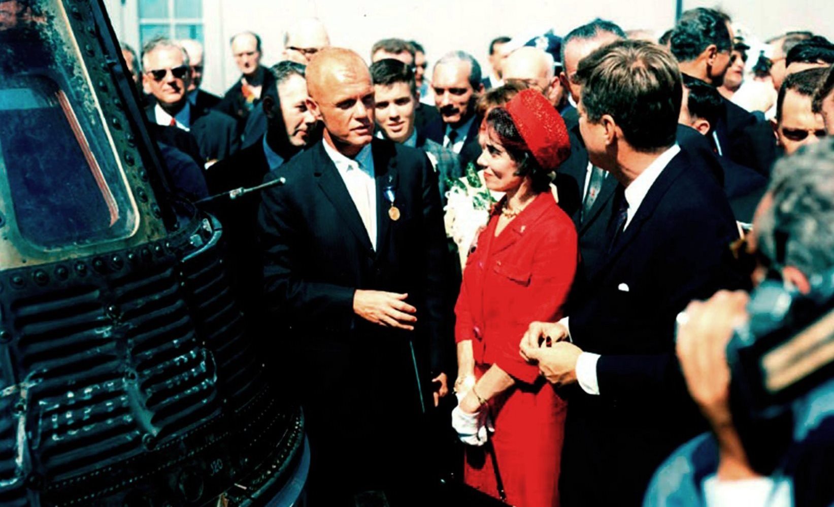 Astronaut John Glenn stands next to the Mercury Friendship 7 spacecraft as he talks to his wife, Annie and President John F. Kennedy. A large group of people surround them.