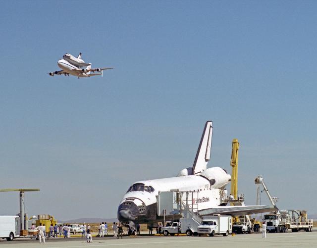 Image from the front cover of NASA Armstrong Flight Research Centers Contributions to the Space Shuttle Program showing a space shuttle parked at AFRC in the foreground and another riding on a shuttle carrier in the sky behind it.