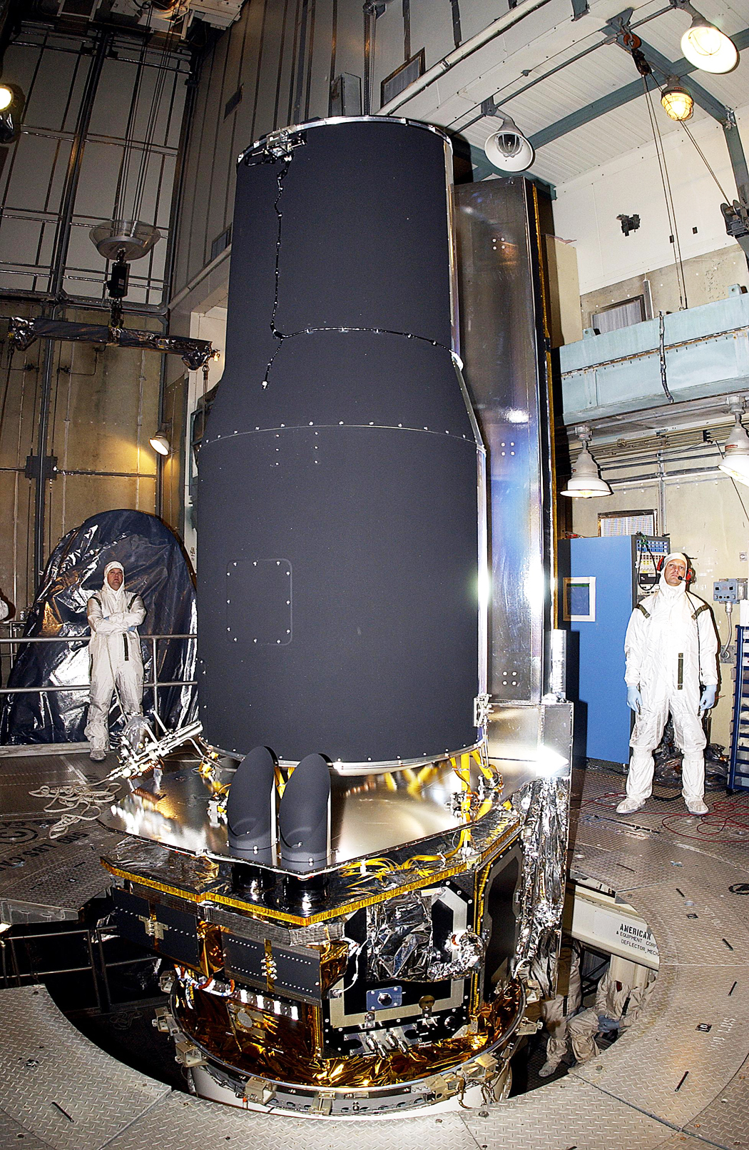 The Spitzer Space Telescope is prepared for launch with a couple of workers looking on.
