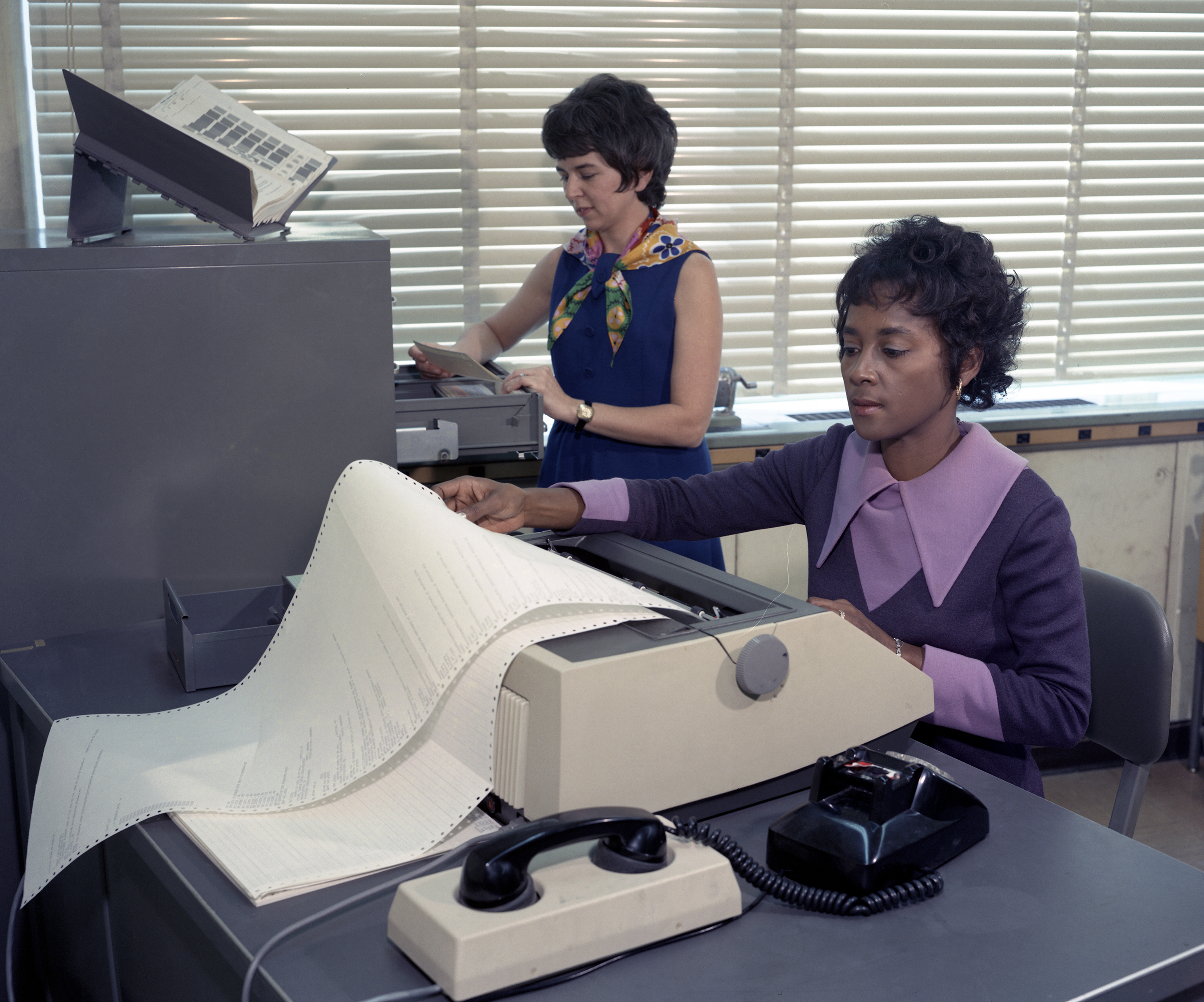 Annie Easley works at a desk with a machine creating a printout while a woman stands at a filing cabinet behind her.