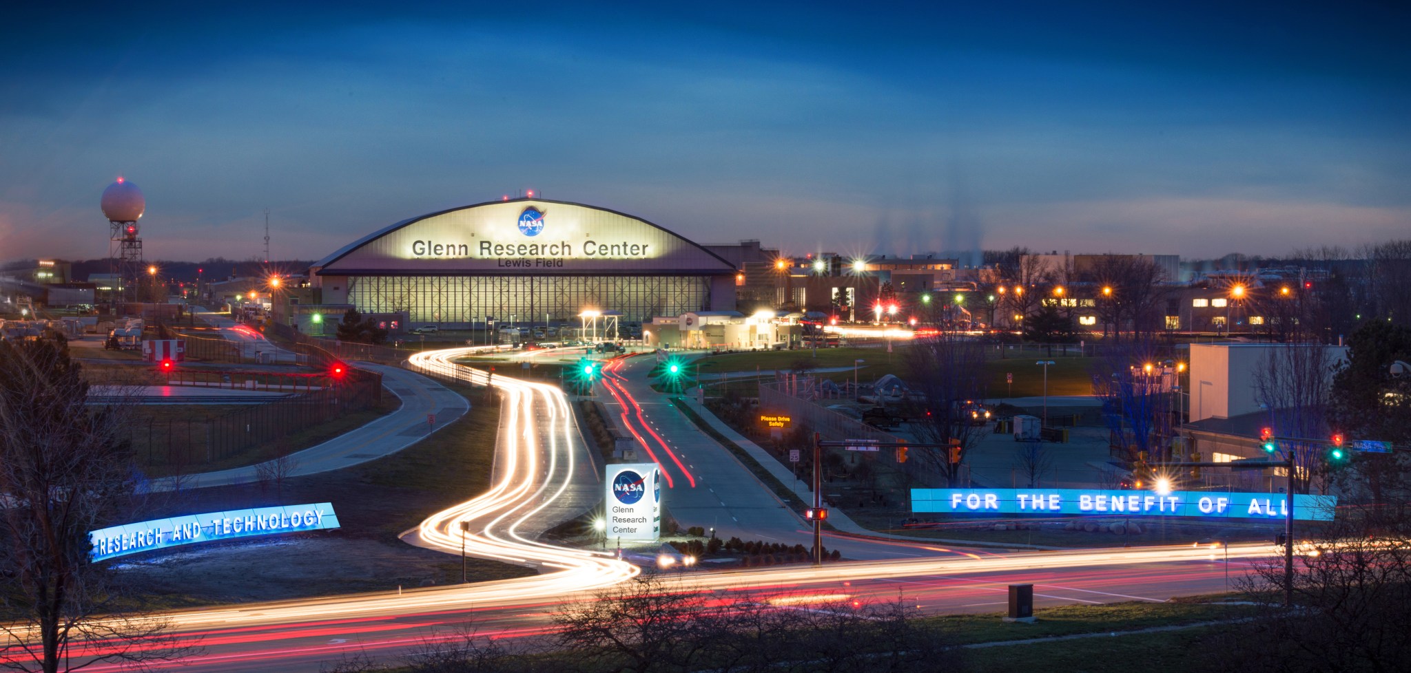 Glenn Research Center Entry at night with lighted hangar in the background featuring the NASA meatball. Signs at the front drive say for the benefit of all.