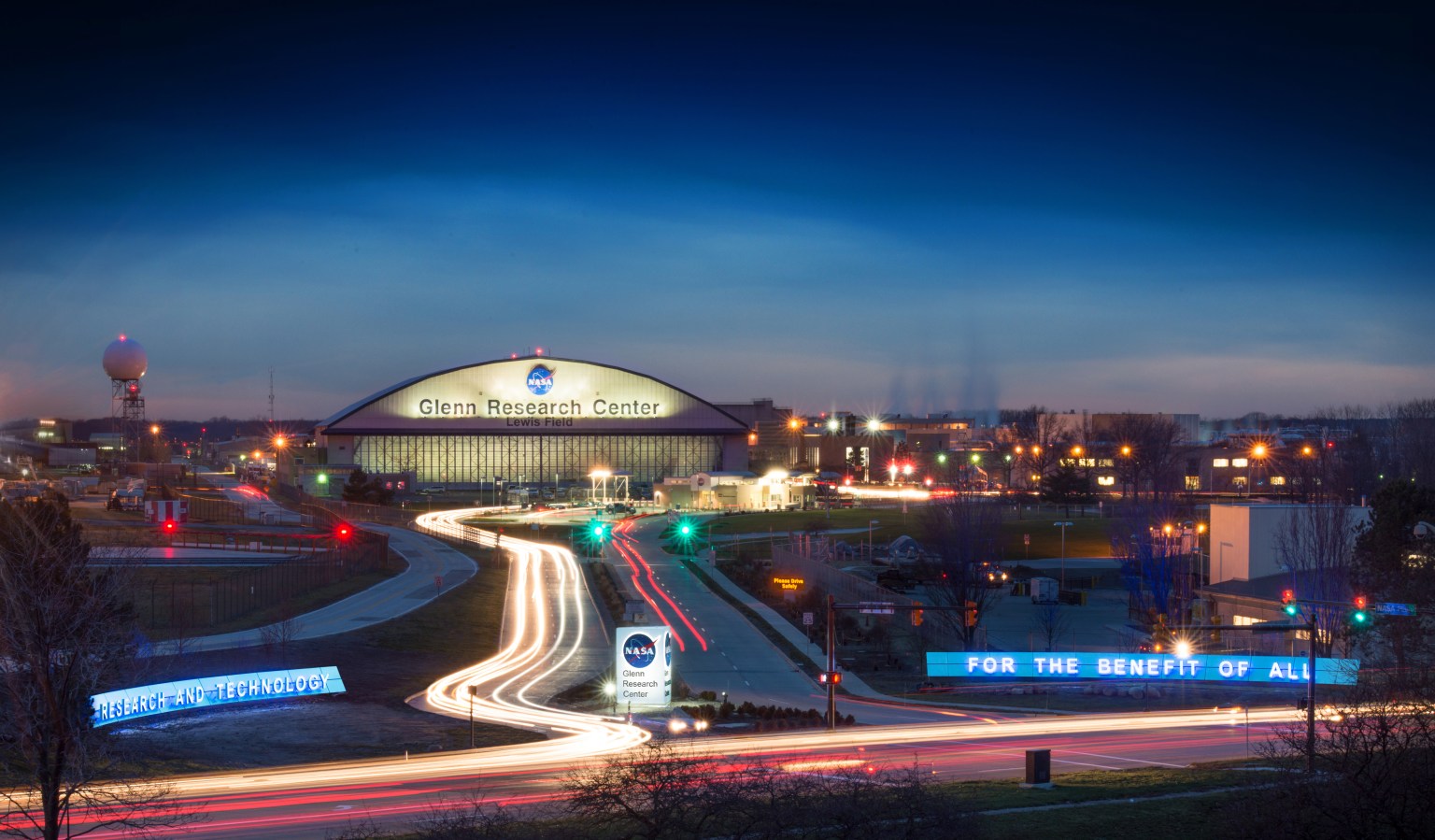 Entry gate to NASA's Glenn Research Center at night with driveway leading to the iconic hangar with the NASA meatball logo on the front.