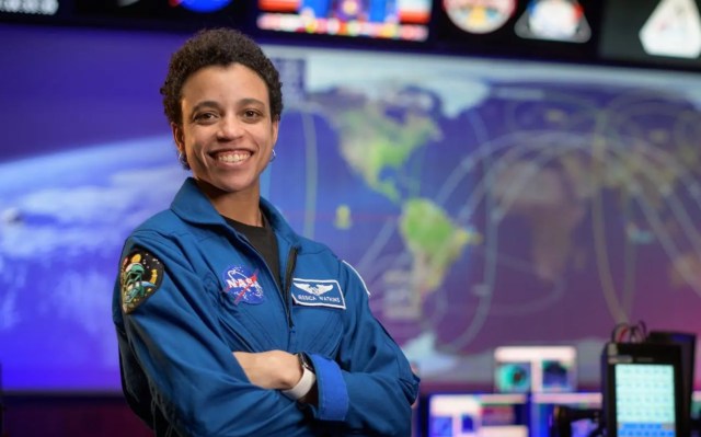 Astronaut Jessica Watkins with crossed arms smiling towards the camera, standing in front of the Mission Control Screens.