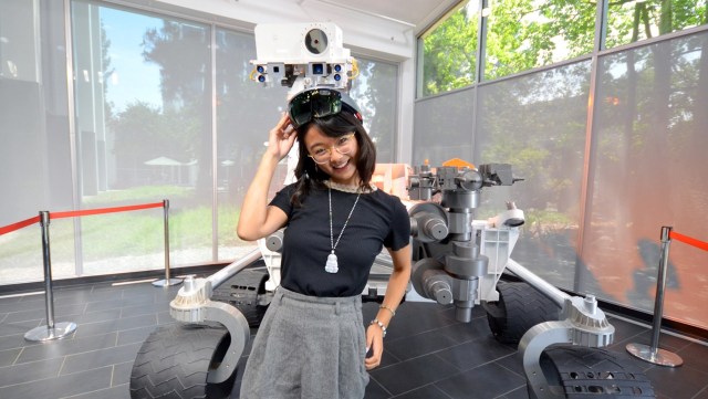 Female holding virtual reality goggles on top of her head, smiling towards the camera, standing in front of a space rover.