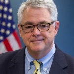 Dave Pierce, a white man with white hair, wears a pair of black-rimmed glasses, a dark-colored blazer with a light blue shirt and yellow tie. Behind him is the American flag against a blue background.