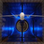 A scale model of an airplane with long and skinny wings supported by a truss is suspended within a wind tunnel for testing. The four walls of the wind tunnel are silver with a blue light illuminating the background.