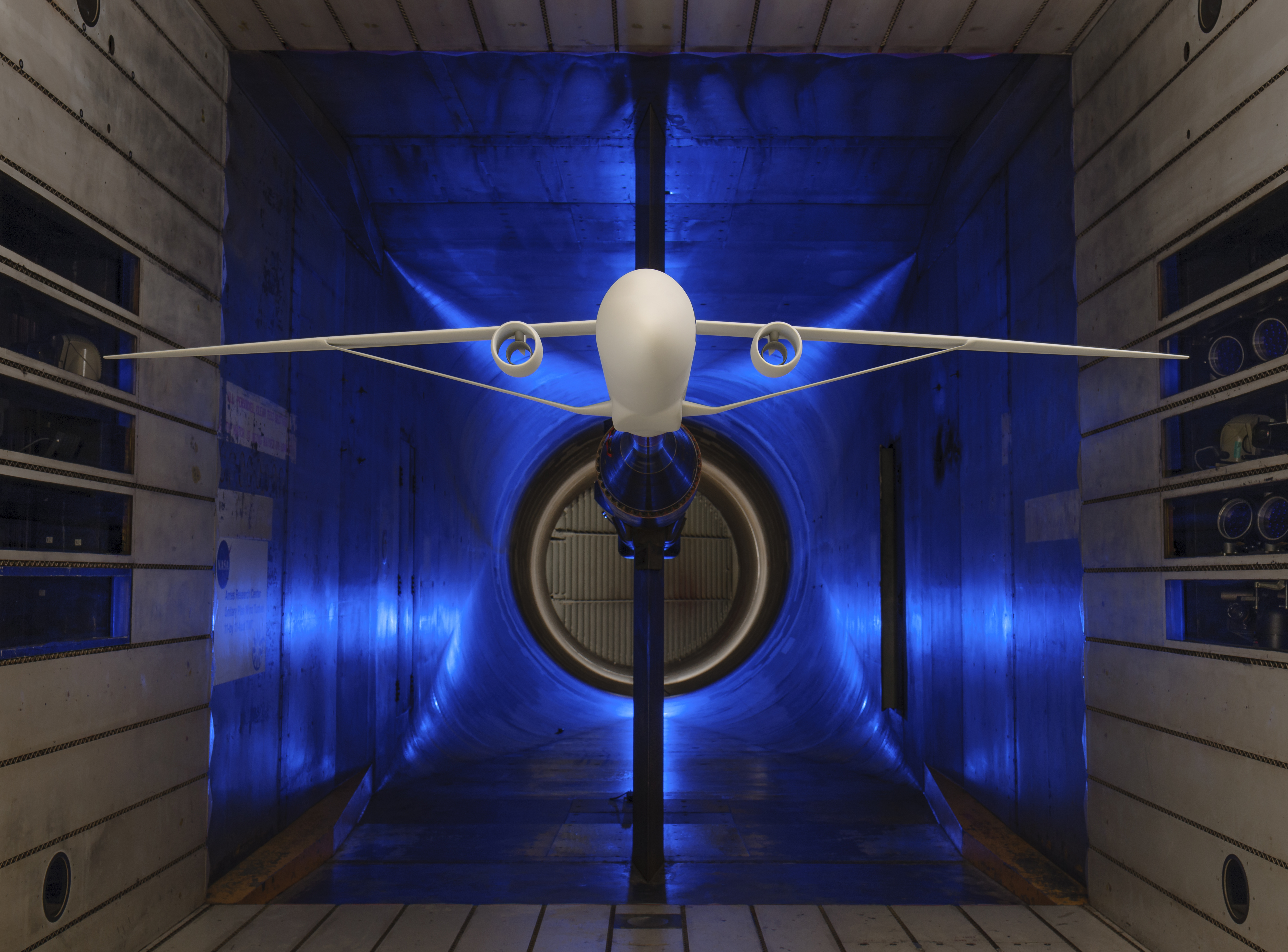 A scale model of an airplane with long and skinny wings supported by a truss is suspended within a wind tunnel for testing. The four walls of the wind tunnel are silver with a blue light illuminating the background.