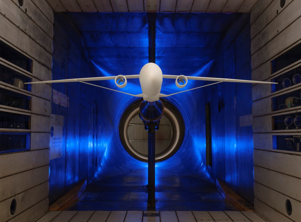 A scale model of an airplane with long and skinny wings supported by a truss is suspended within a wind tunnel for testing. The four walls of the wind tunnel are silver with a blue light illuminating the background.