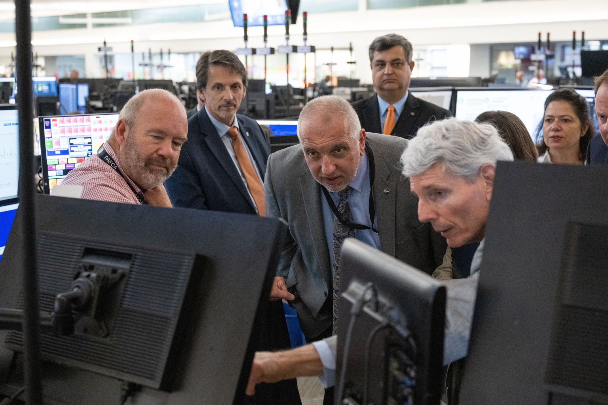 A group of people gather in front of monitors. One person is speaking and pointing at a monitor.