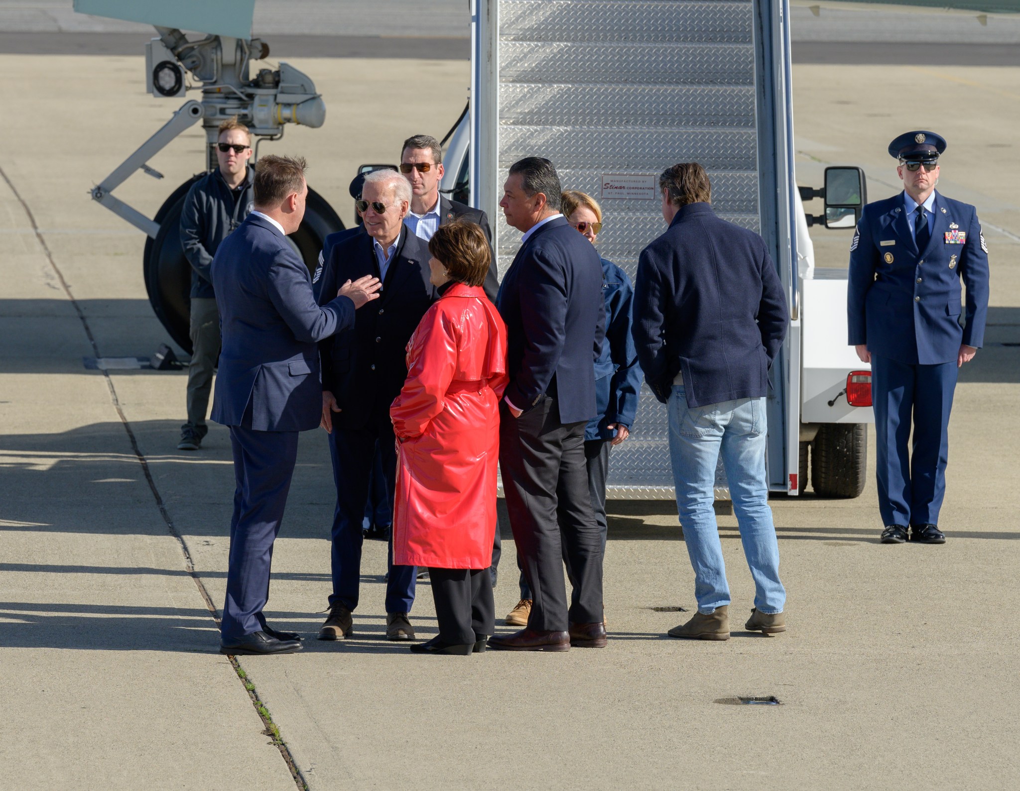 A group of people gather in front of the stairs to an airplane. At center, President Joe Biden speaks to two men in dark blue suits and a woman in a red raincoat.