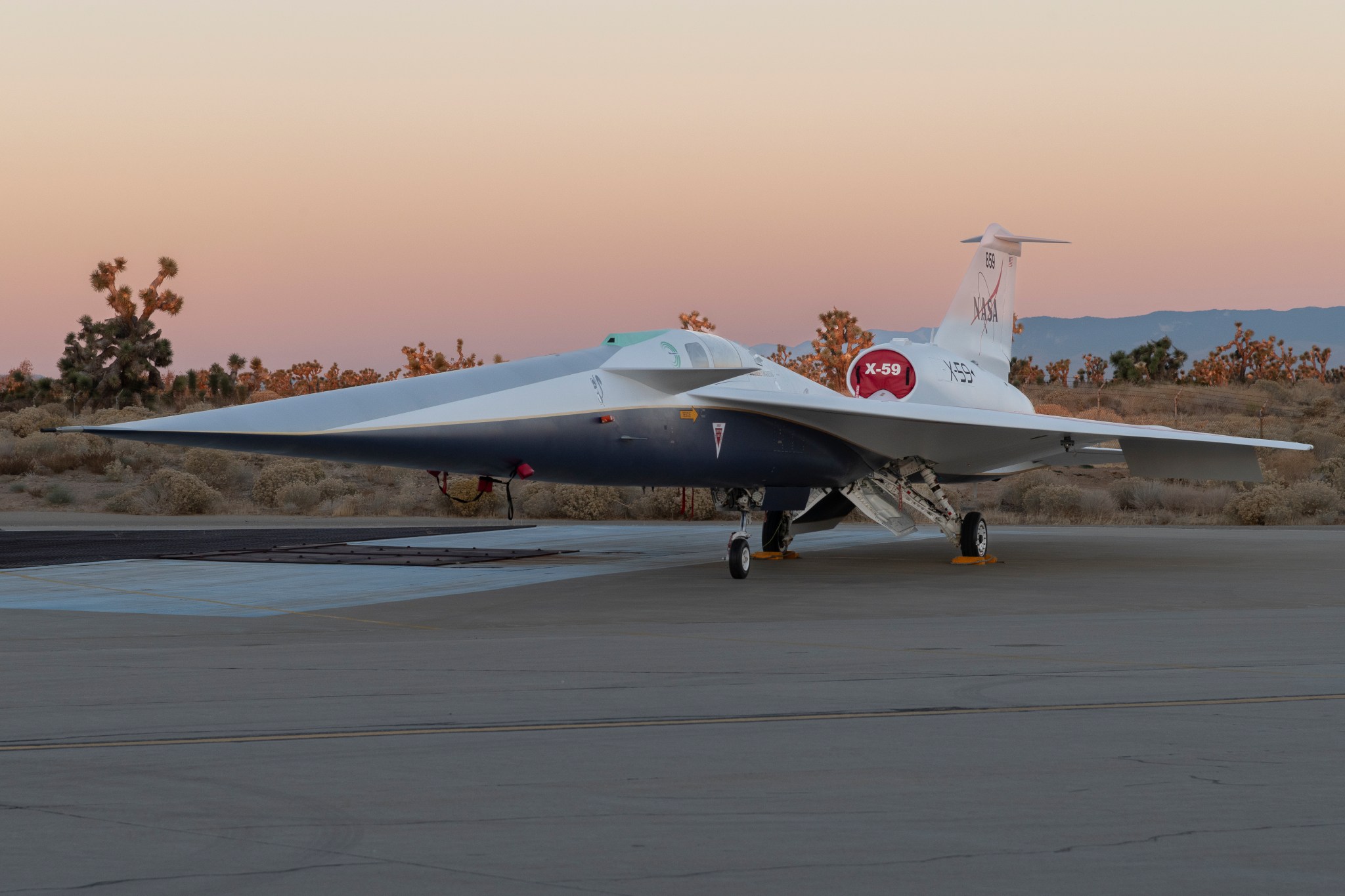 In this photo, NASA’s X-59 aircraft is shown at sunrise at the Lockheed Martin Skunk Works in Palmdale, California. Open desert can be seen behind the aircraft. The X-59’s design features a long nose, approximately one-third of the aircraft’s nearly 100-foot length, swept-back wings, and an engine mounted on top and toward the rear of the fuselage.