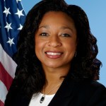 Christyl Johnson with long, flowing black hair. She is wearing a black blazer with a white shirt underneath. The background is blue with an American flag over her right shoulder.