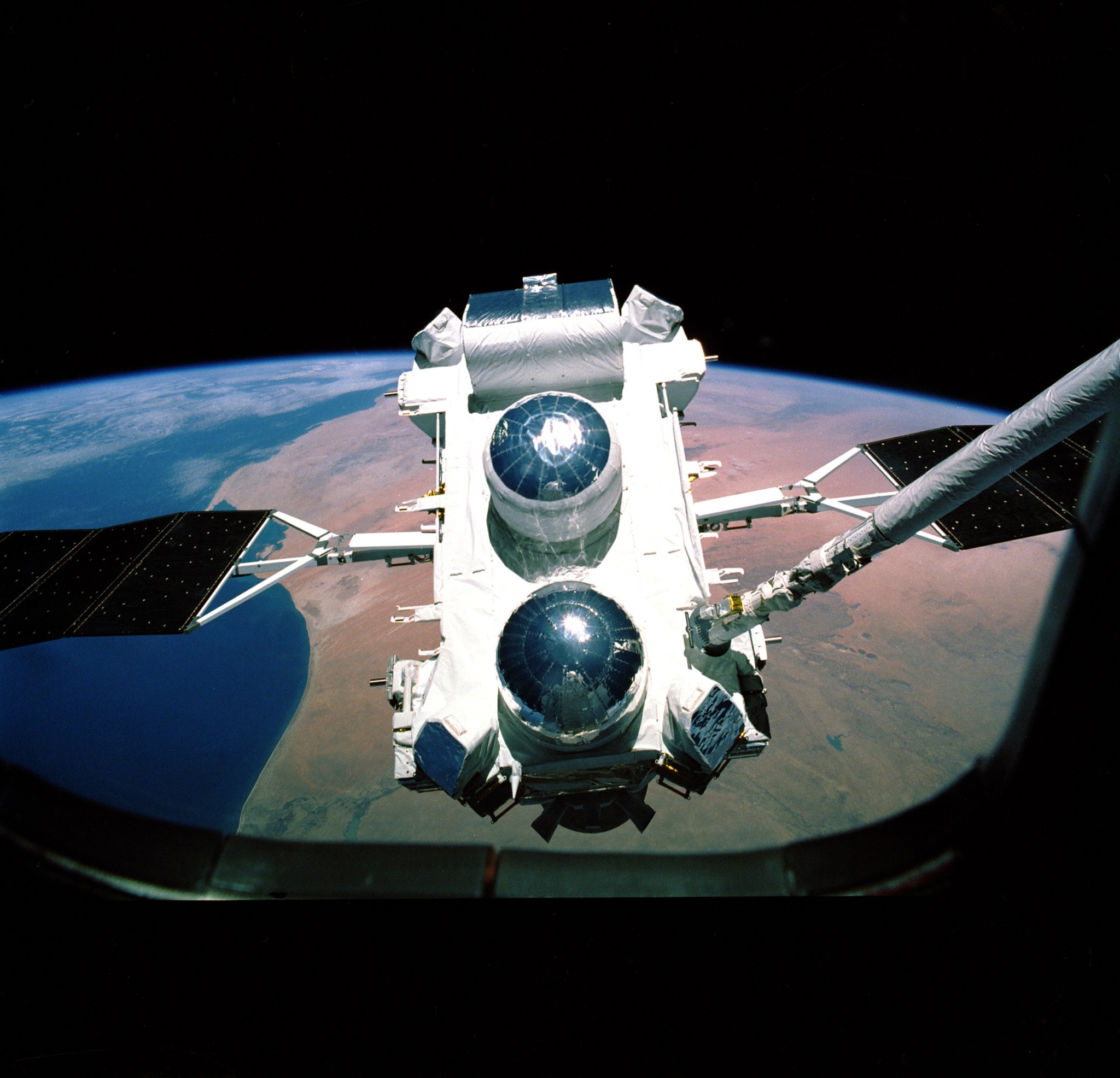 A white, rectangular spacecraft floats above a tan and blue Earth outside a window on space shuttle Atlantis. The spacecraft has two silvery domes at the near end a silver cylindrical element at its far end. Two dark solar panels extend left and right.
