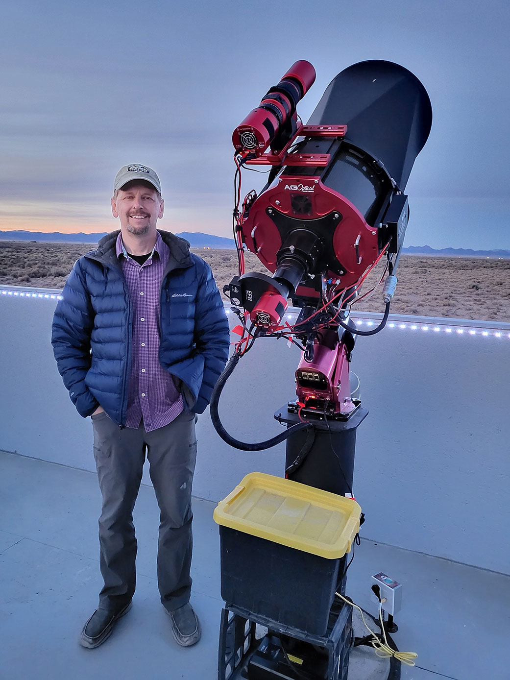 Photograph of a person stands on a concrete patio wearing a hat and a blue coat, next to a black and red telescope. In the background is a desert landscape, with mountains in the distance.