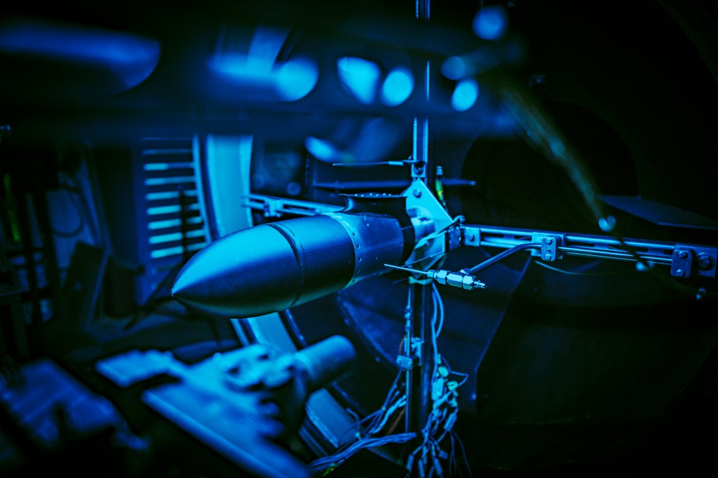An aluminum section behind a nose cone inside a blue-lit wind tunnel during testing.