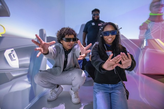 Two students in virtual reality glasses stretch out their hands to interact with what they are seeing on their internal screens.