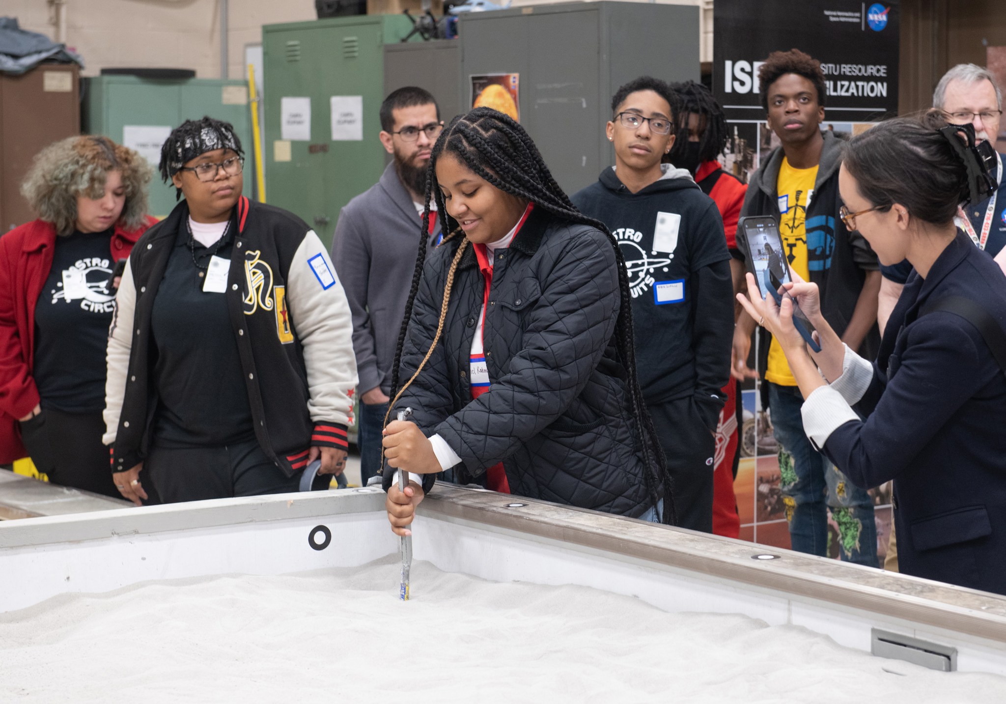 A group of students looks on while a young woman student digs in a sandbox with a metal rod, while the teacher takes a photo on her phone.