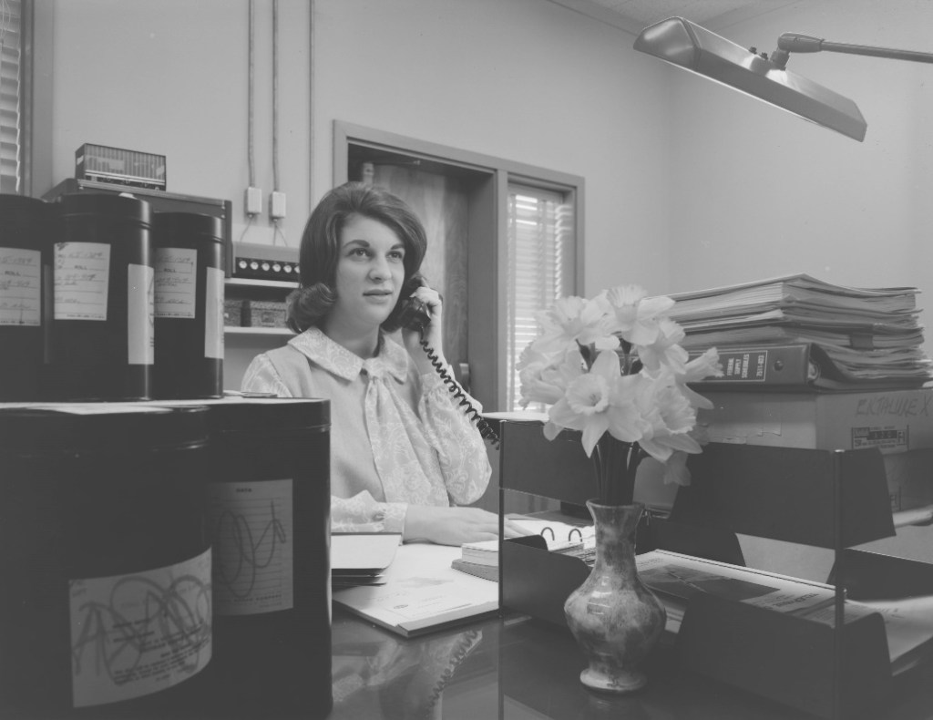 A woman holds a phone receiver to her ear as she works at a desk.