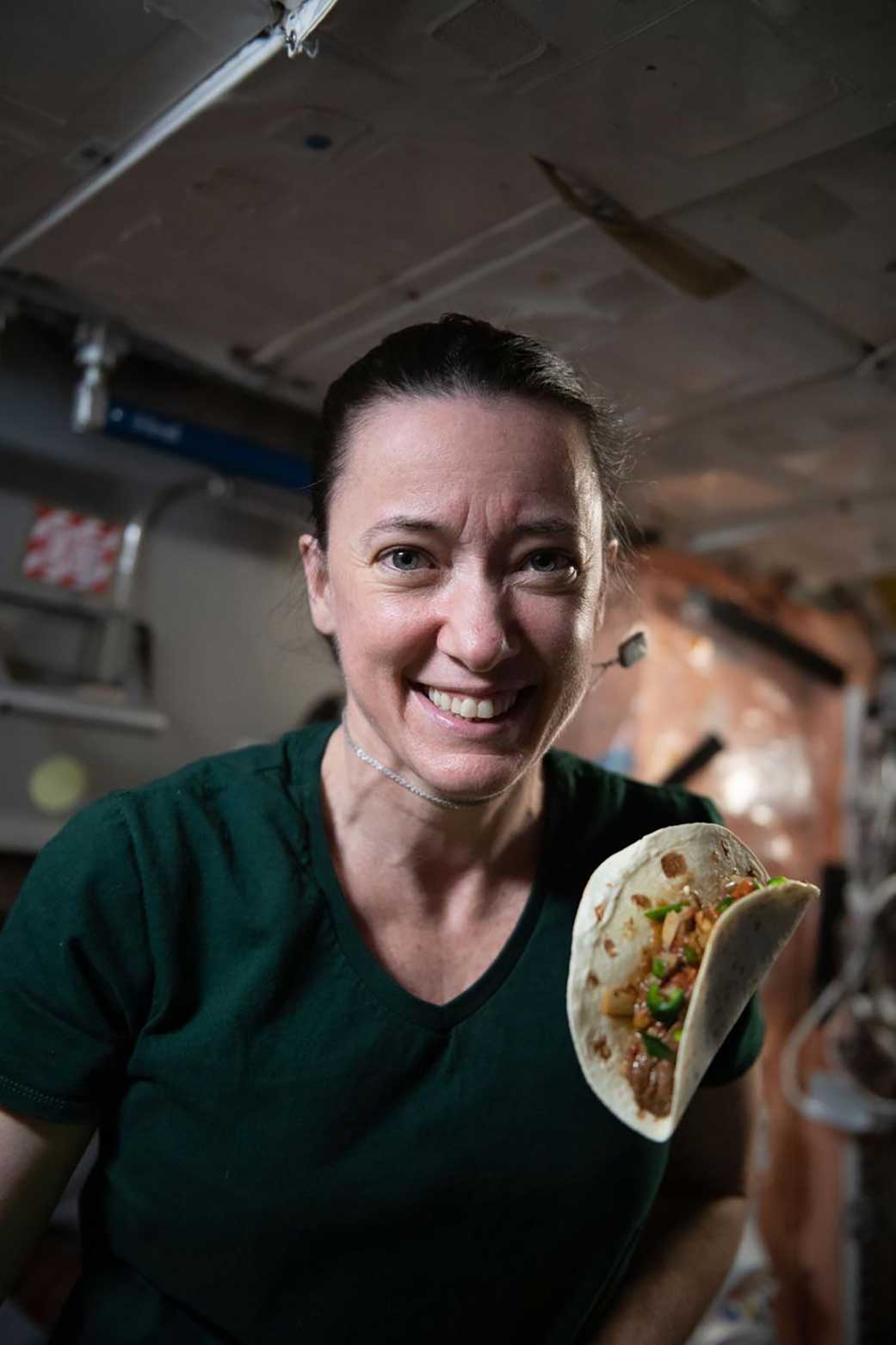 A female astronaut on the iSS wearing a green shirt has a taco floating in front of her.