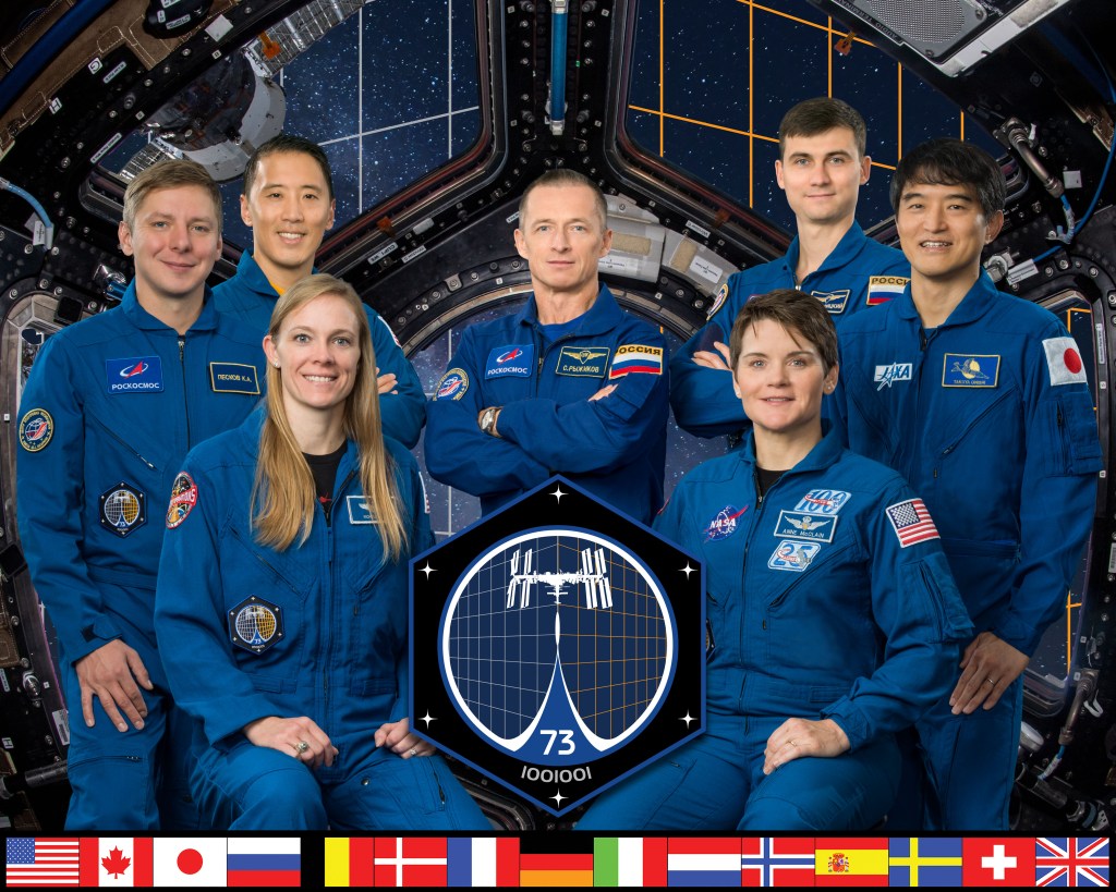 Six people in blue flight suits pose for a picture. The blue, black and white Expedition 73 patch is in the middle of them and the image is bordered on the bottom by the country flags of the nations who helped created and still support the space station.