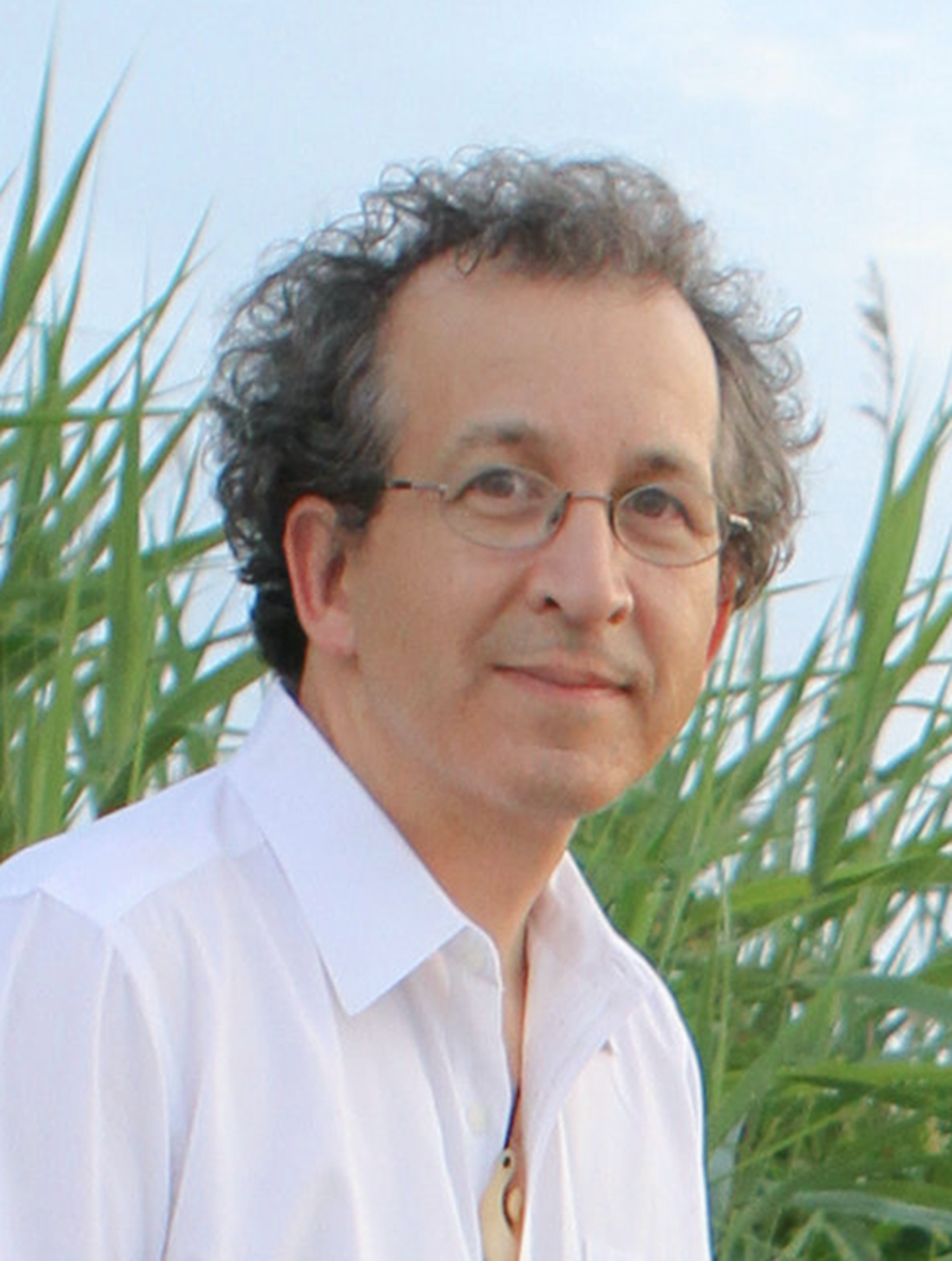 A white man with curly gray hair and glasses smiles slightly in front of a backdrop of green sea grasses and blue sky. He wears a white shirt and necklace.