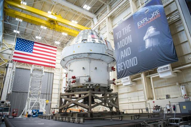 A view of the Orion Spacecraft as it sits inside the Space Environments Complex high bay. A large American flag is hung on the wall along with a poster that reads, "To the Moon and Beyond."