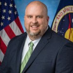 A bearded, bald man in a suit and tie poses in front of the American and NASA flags