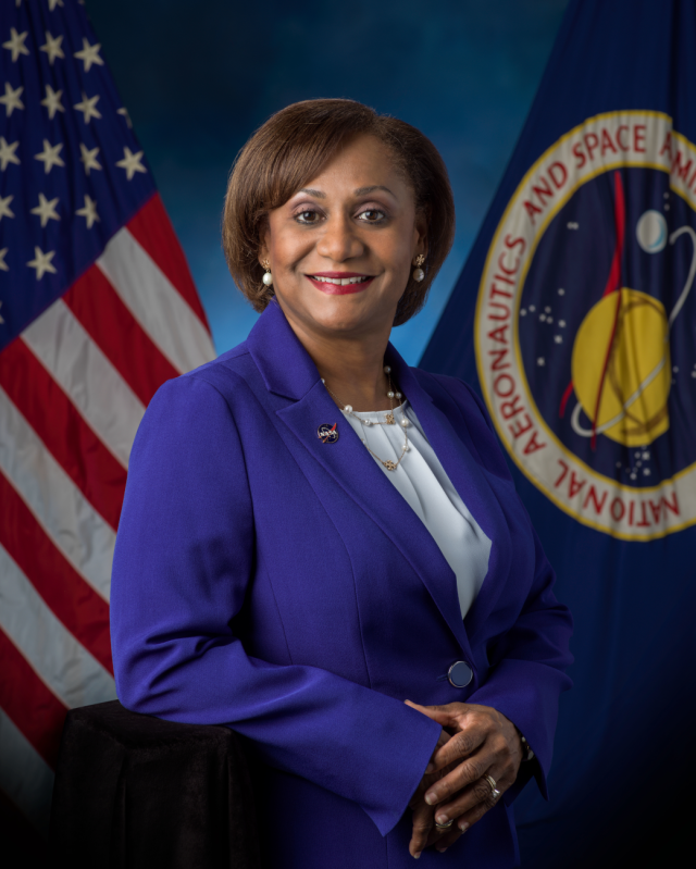 Vanessa Wyche poses in front of the United States and NASA flags wearing a blue suit and white top.