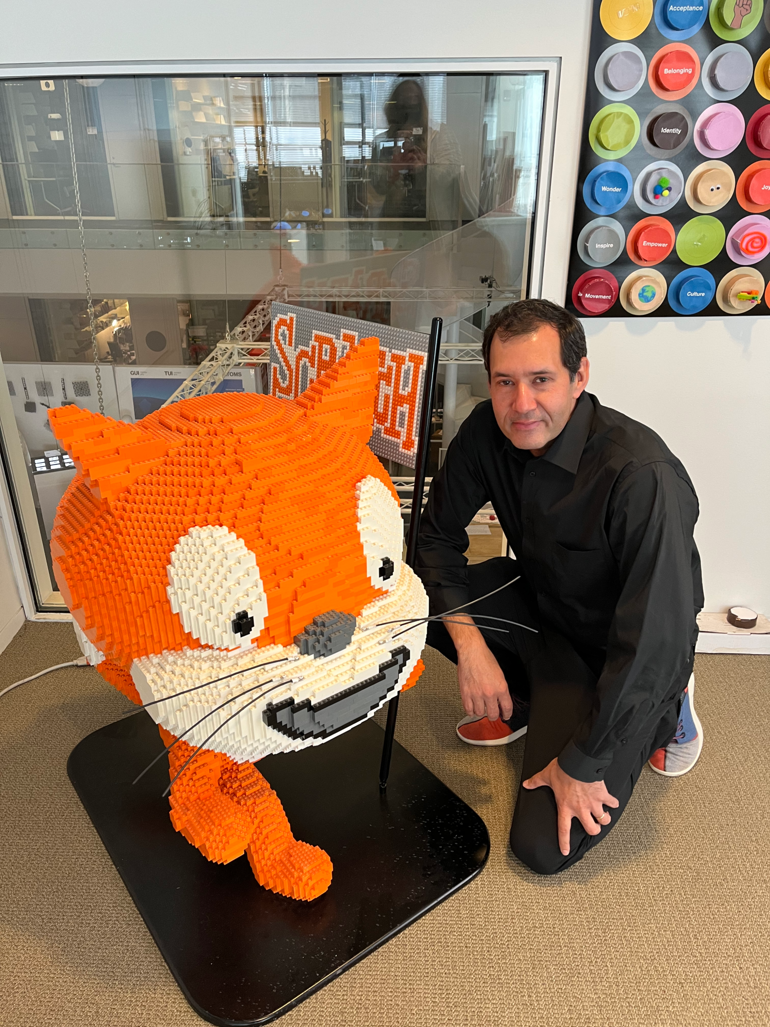 A man with dark hair kneels next to a large LEGO sculpture of an orange and white cartoon cat.