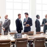 Several people in business suits appear talking as they stand behind an office conference room table. On a nearby wall is a graphic with an airplane and the words NASA Advisory Council Aeronautics Committee.