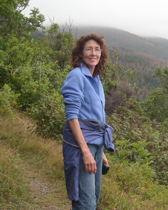 A women with long, curly brown hair, glasses, wearing a blue shirt and jeans poses outdoors with a hilly, heavily forested area behind her.