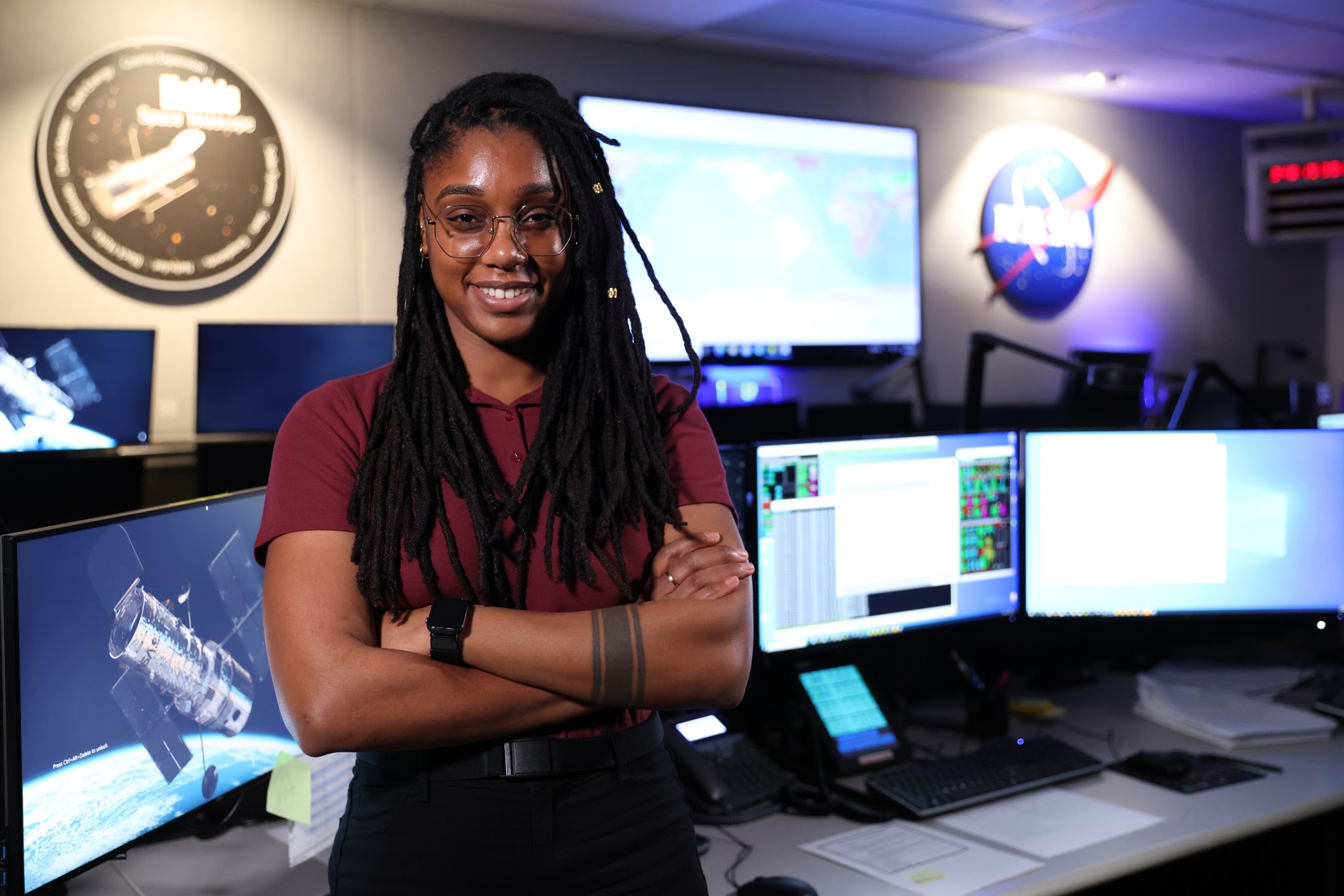 Phanthom Donald, a Black woman with long black dreadlocks and glasses, smiles and poses in the Hubble Space Telescope control room. She wears a burgundy polo and black pants and has a black tattooed band around her left forearm.
