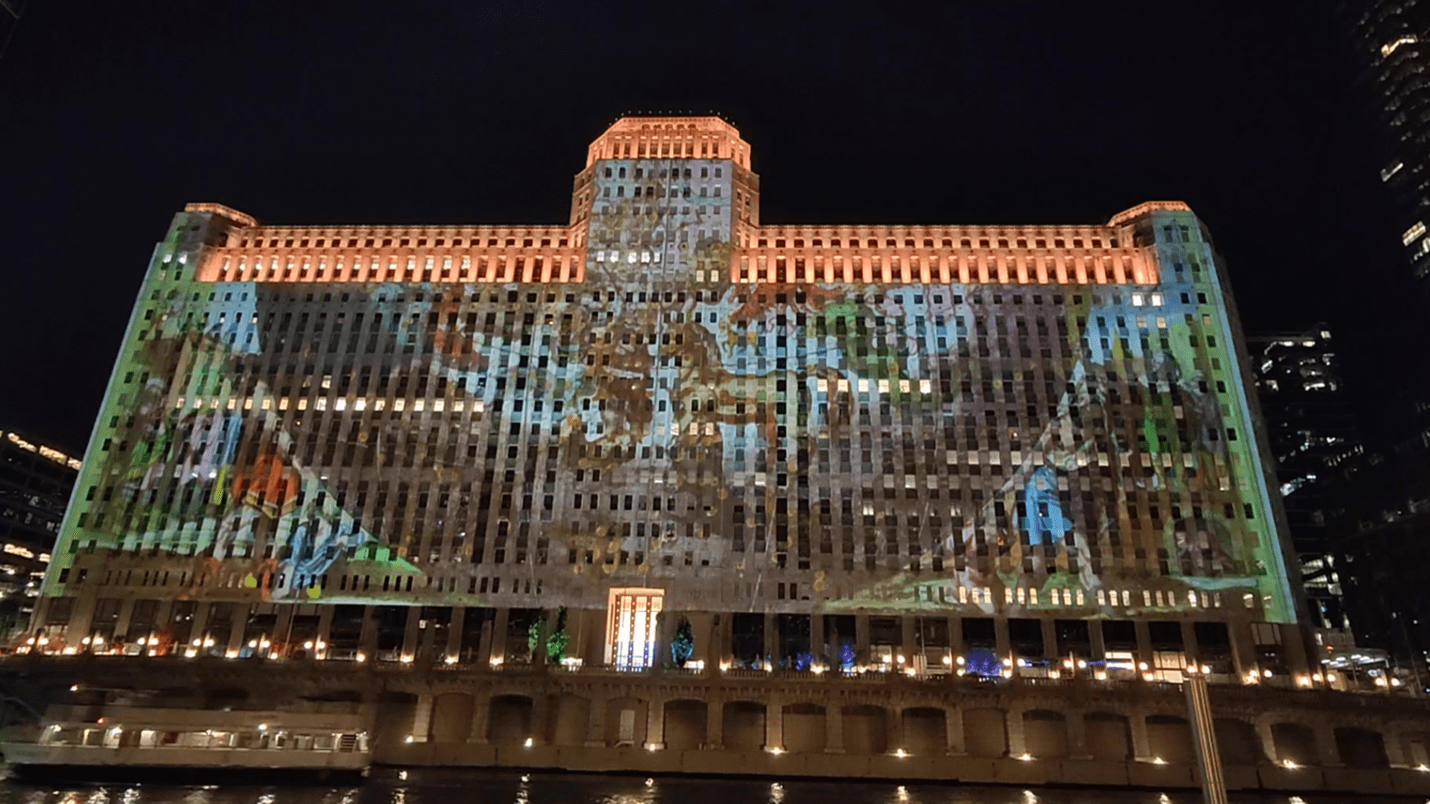 A large building with many windows is covered by a projection of an art piece. The art shows a globe with a horse and other animals in the center, and figures on either side of the globe.