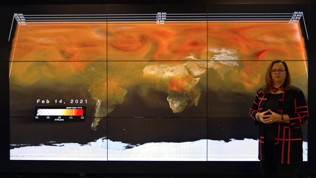 A woman with dark hair and glasses stands in front of a data visualization of carbon dioxide. The visual is a flat map of the Earth, with swirling clouds of orange and red depicting carbon dioxide.