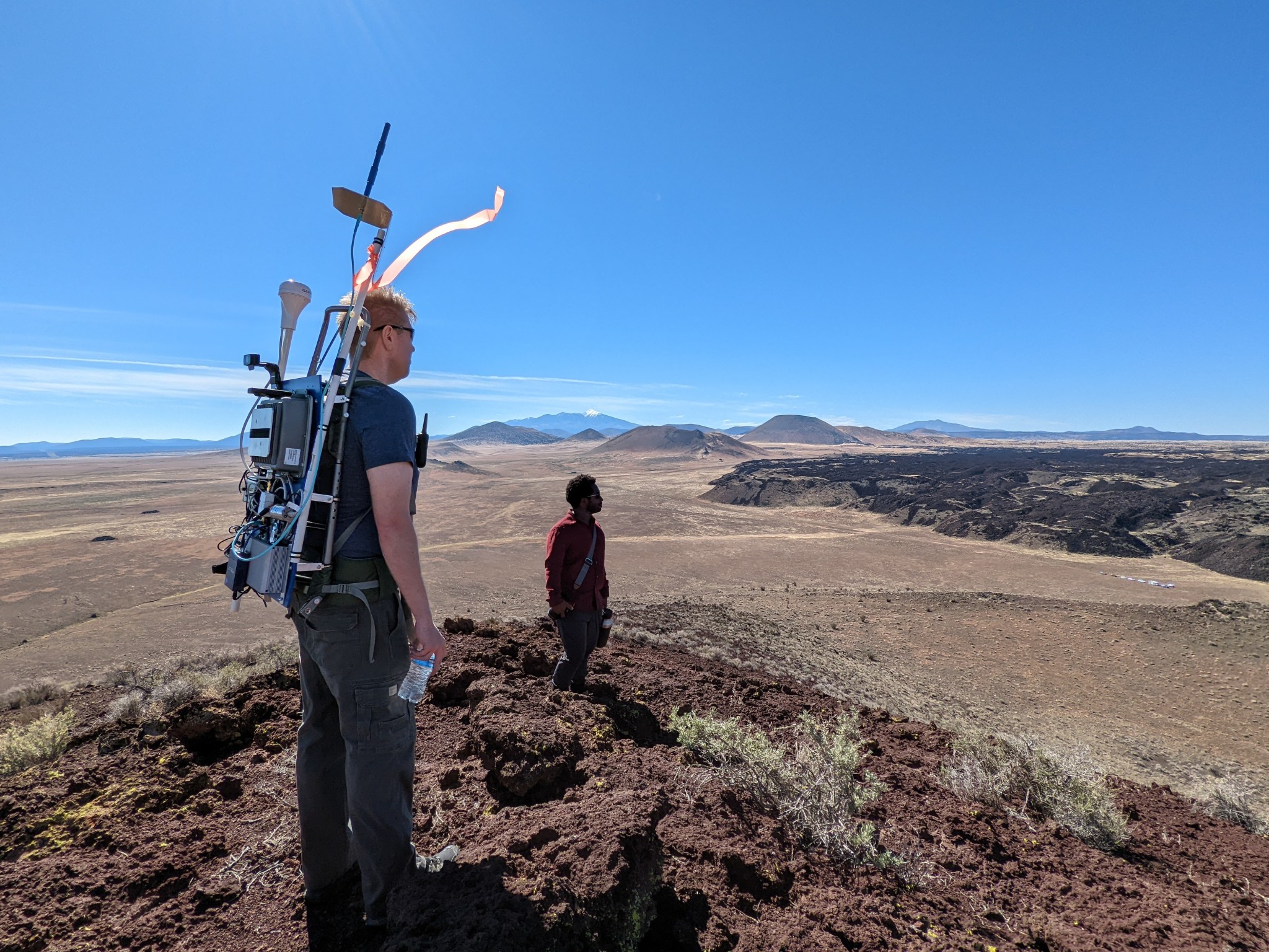Two men standing in the desert, one is in the background nearly in silhouette, and one in the foreground has some test equipment loaded on a back pack. Both look into the distance to the right of the frame. Blue sky and mountains in the distance.