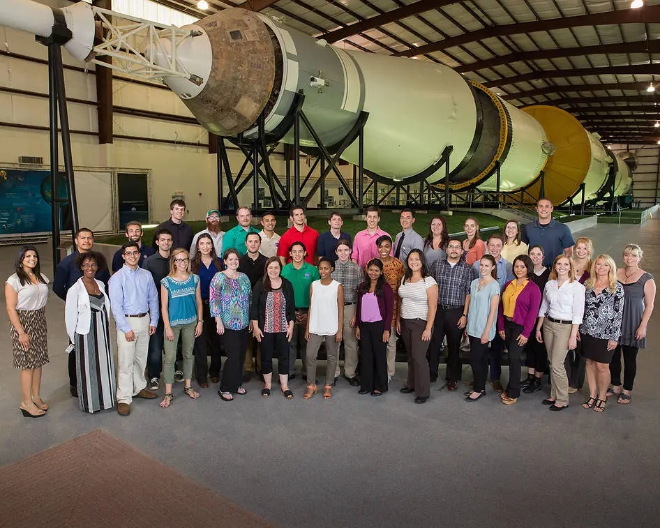 A large group posing and smiling in front of the camera, standing in front of a rocket display.